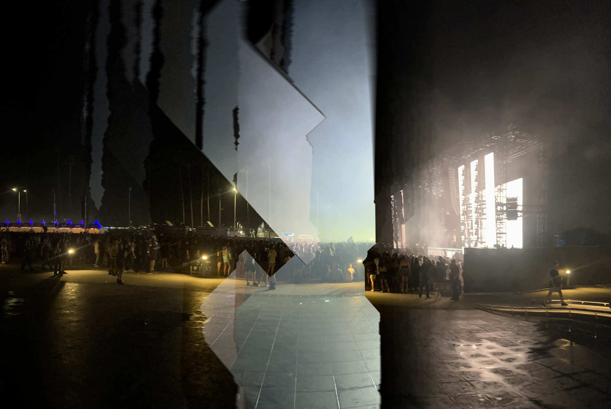 A festival stage at night, captured mid-panorama, the left half torn into vertical bands while the stage blows out to white on the right.